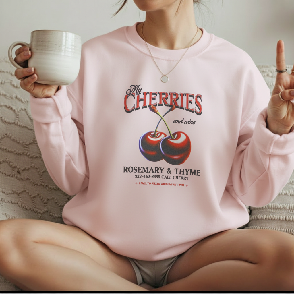 Woman wearing light pink My Cherries vintage graphic sweatshirt sitting on bed with coffee mug
