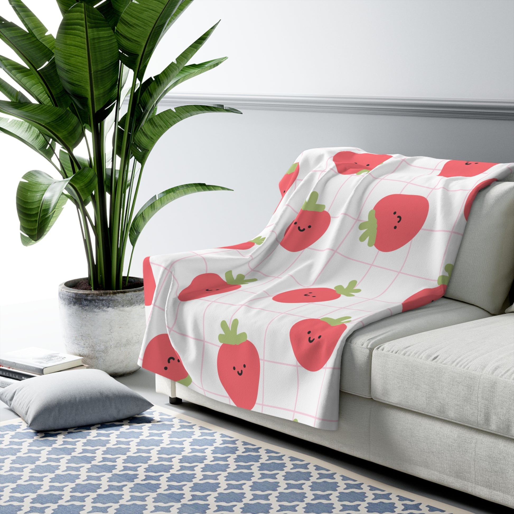 White sofa with a strawberry-patterned blanket, plant, and rug in a room.